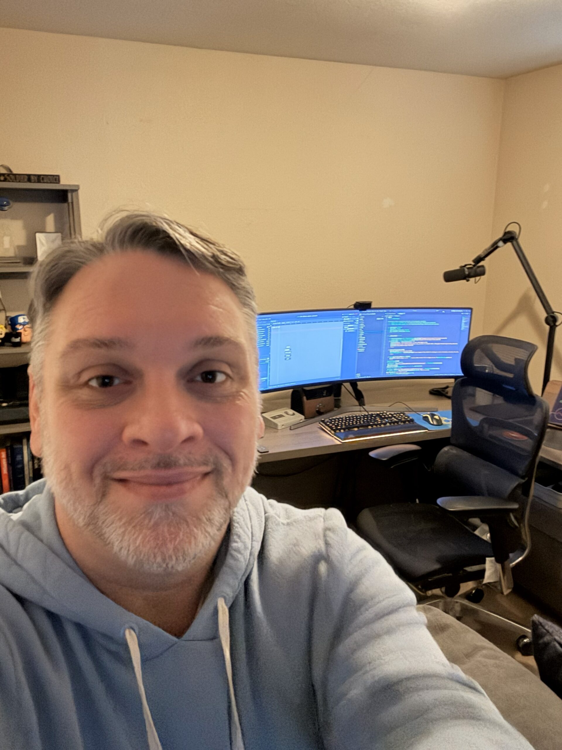 A headshot of Tim Moore smiling in his home office, with a desk featuring a curved ultrawide monitor displaying code, a mechanical keyboard, and a microphone in the background.